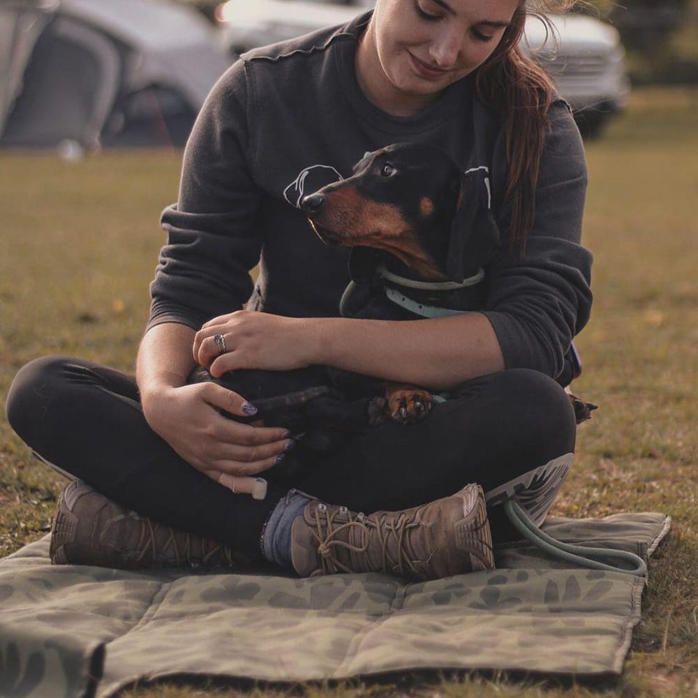 Someone sat on the settle mat with their sausage dog. Taylor and Floof Jungle Pup settle mat featuring a dark green camo print with illustrated dogs, ideal for puppy training and assistance dog training.